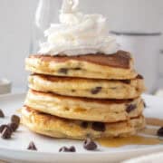 A stack of flour fluffy chocolate chip pancakes topped with syrup and whipped cream on a white plate.