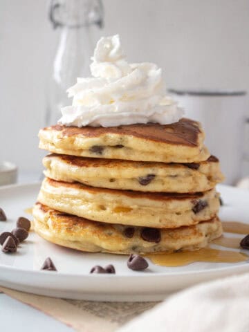 A stack of flour fluffy chocolate chip pancakes topped with syrup and whipped cream on a white plate.