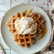 Three cocoa buttermilk waffles on a plate topped with whipped cream and a drizzle of syrup surrounded by heart sprinkles and a rose napkin.