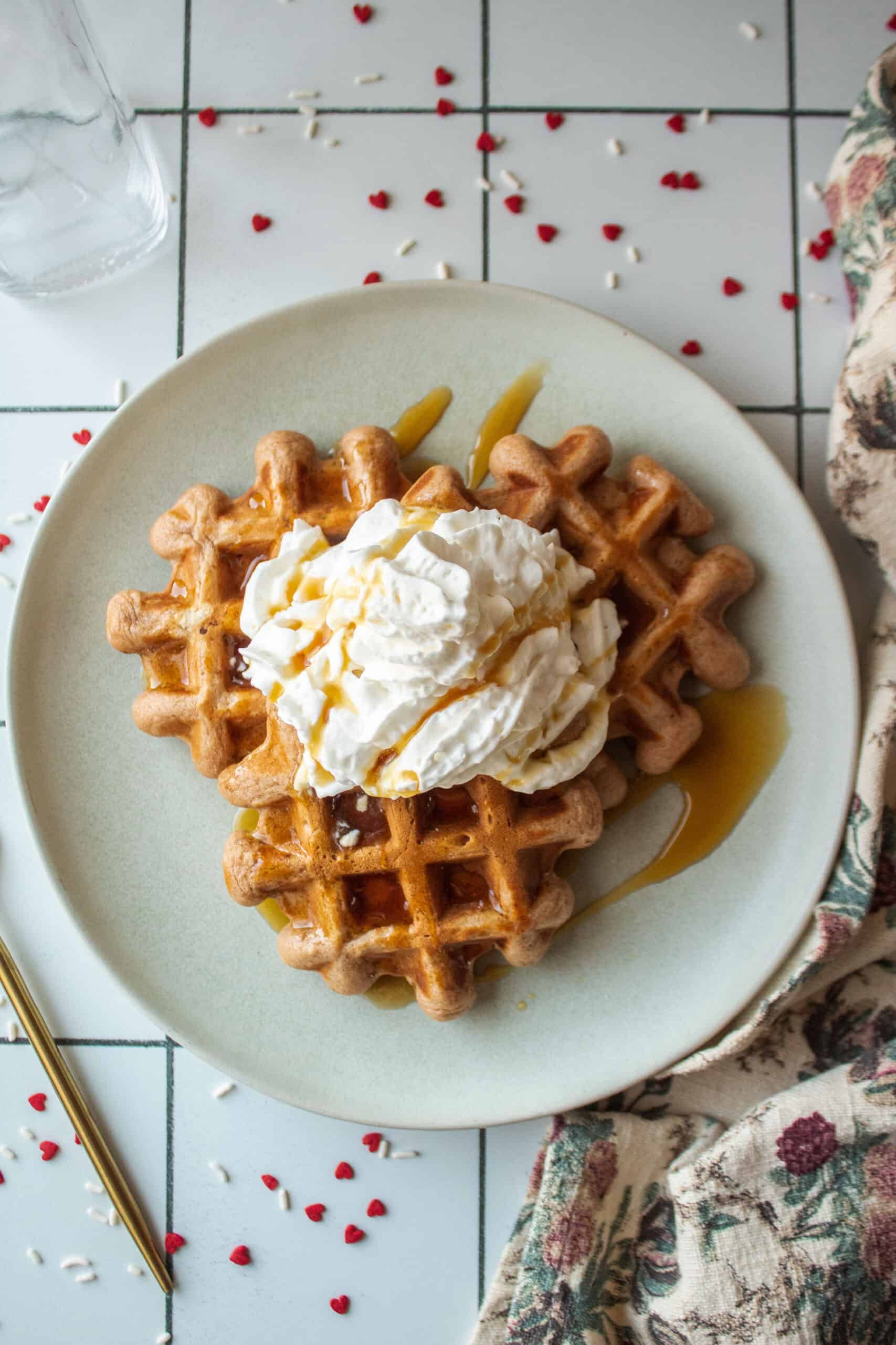 Three cocoa buttermilk waffles on a plate topped with whipped cream and a drizzle of syrup surrounded by heart sprinkles and a rose napkin.