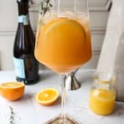 a glass of an orange cocktail with orange slices and a bottle of champagne in the background