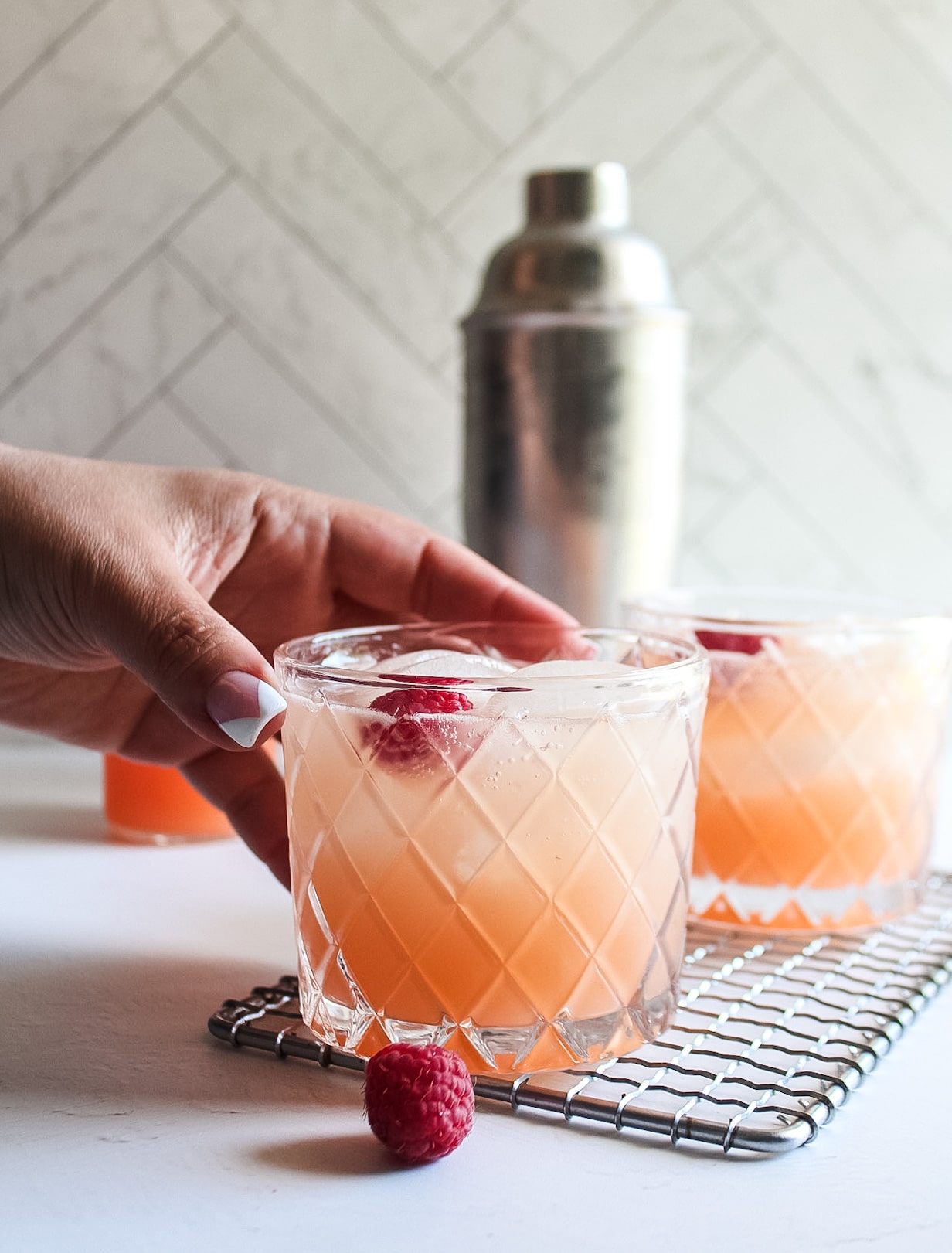 a grapefruit cocktail in a glass with a shaker in the back, a hand reaching in to grab the drink