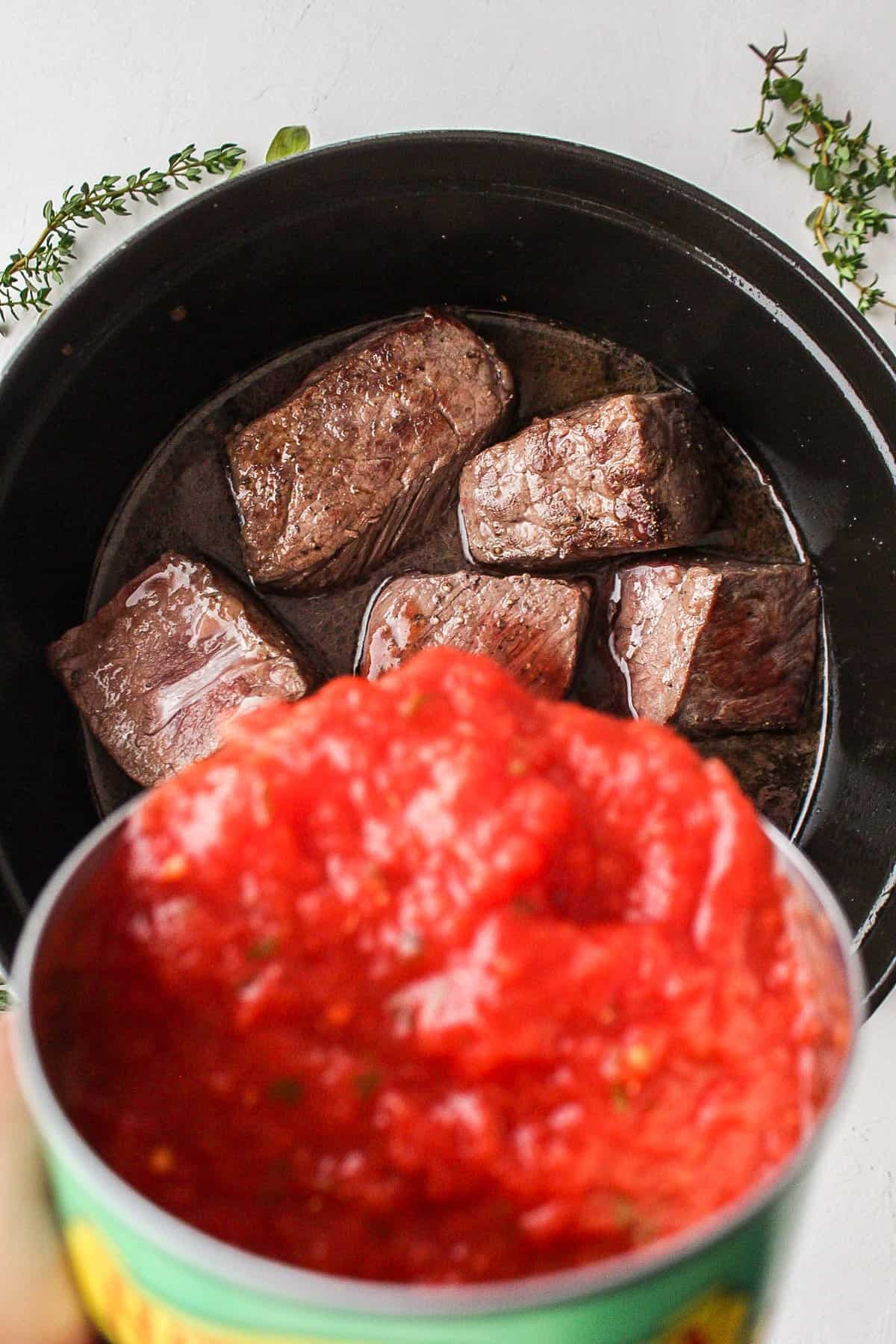 pouring a can of crushed tomatoes into a dutch oven with boneless short ribs