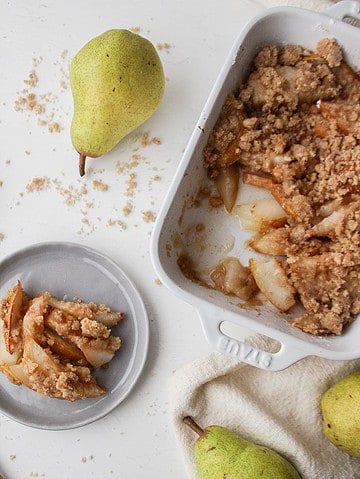pear crumble in a baking dish with some crumble on a plate surrounded by pears