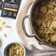 matzo ball chicken noodle soup in a large pot with a mug of soup on the side and a packet of bone broth powder.