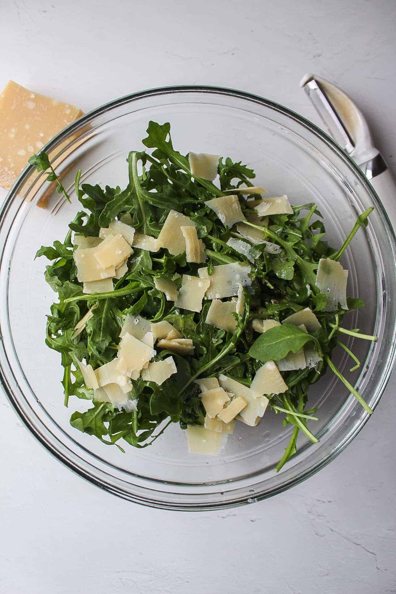 arugula lettuce in a large bowl with fresh shaved pieces of parmesan cheese on top.