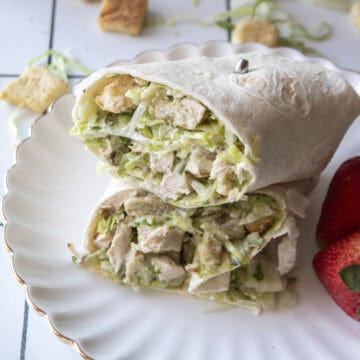 A chicken caesar salad wrap stacked on top of one another on a scalloped white plate with strawberries next to it.