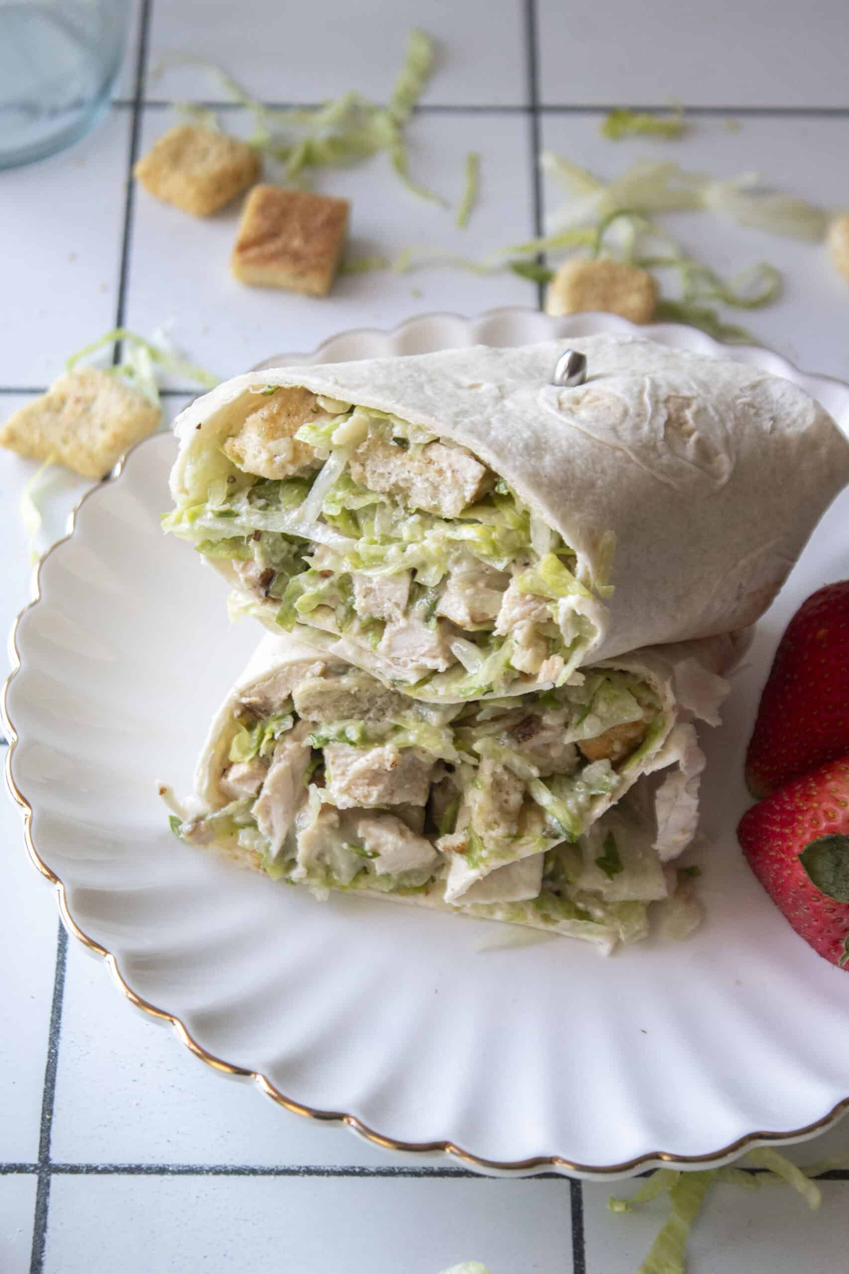 A chicken caesar salad wrap stacked on top of one another on a scalloped white plate with strawberries next to it.