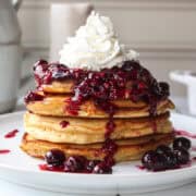 a stack of four almond butter pancakes with a berry compote topped with whipped cream on a plate.