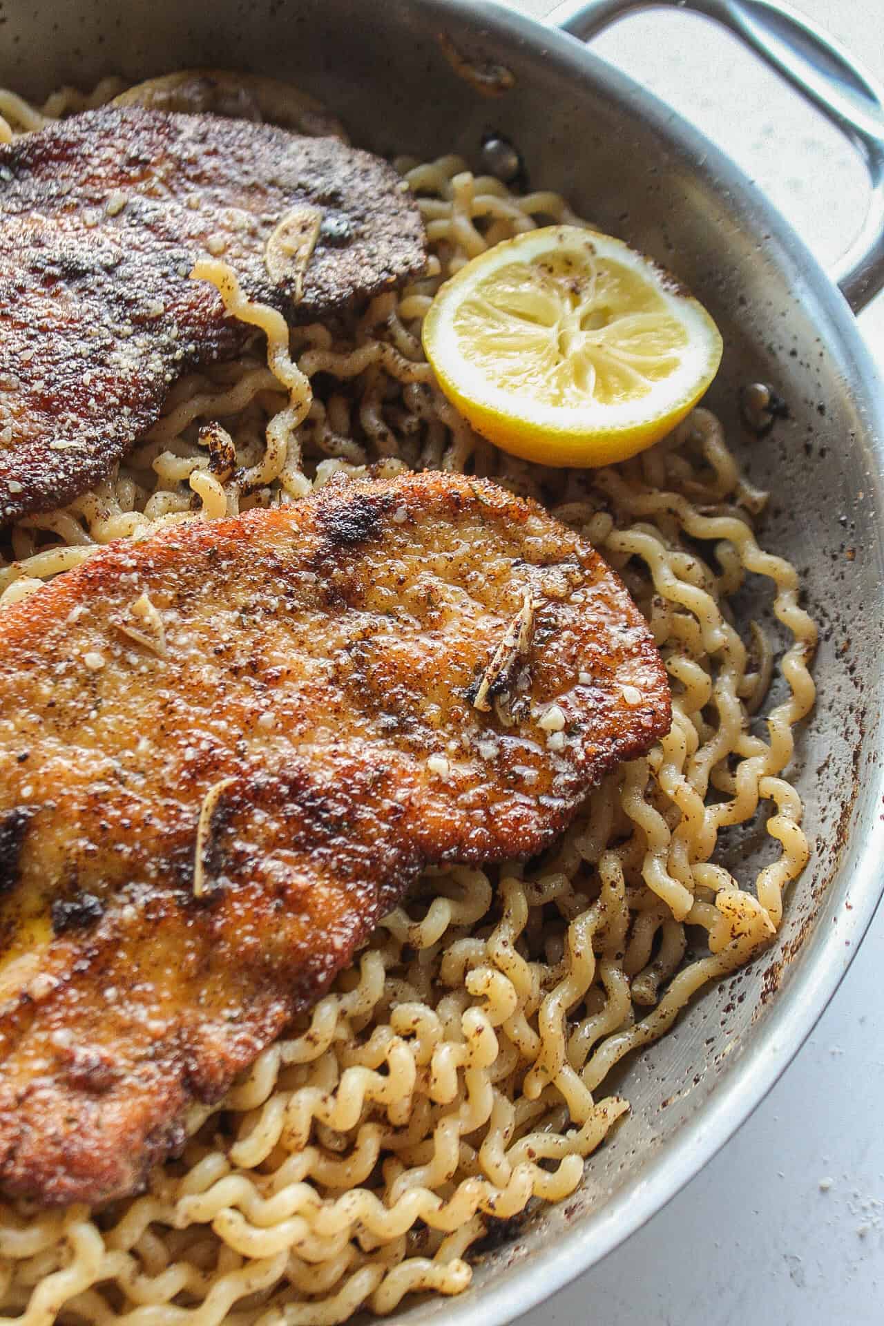 Close up of a chicken cutlet and lemon slice over pasta in a skillet.