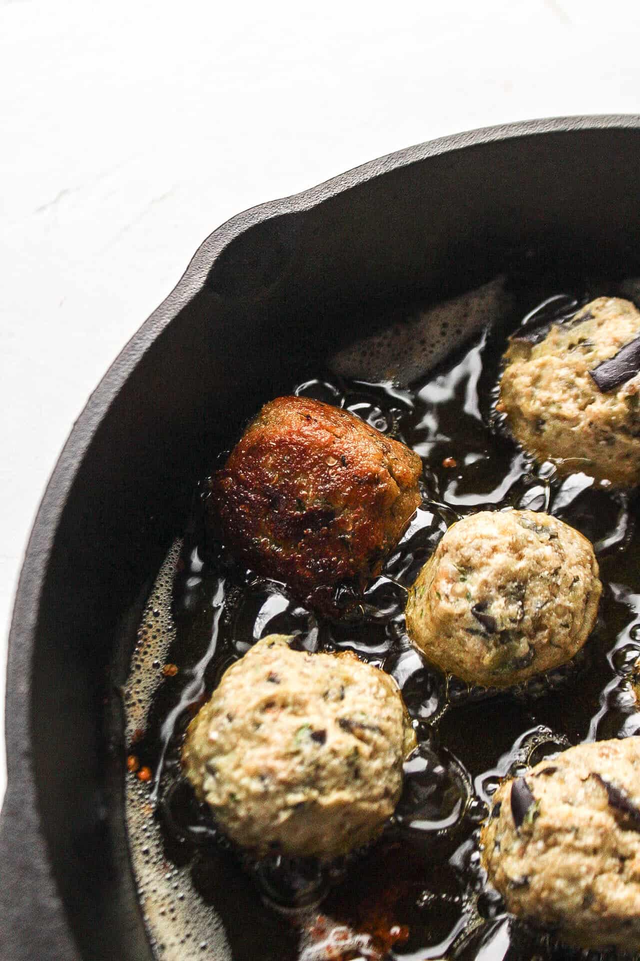 Eggplant meatballs frying in a cast iron skillet with oil.