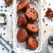 Six chocolate protein muffins in a porcelain basket with chocolate chips surrounding.