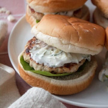 greek chicken burger on a plate topped with tzatziki with the top bun sliding off.
