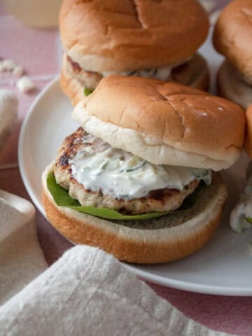 greek chicken burger on a plate topped with tzatziki with the top bun sliding off.