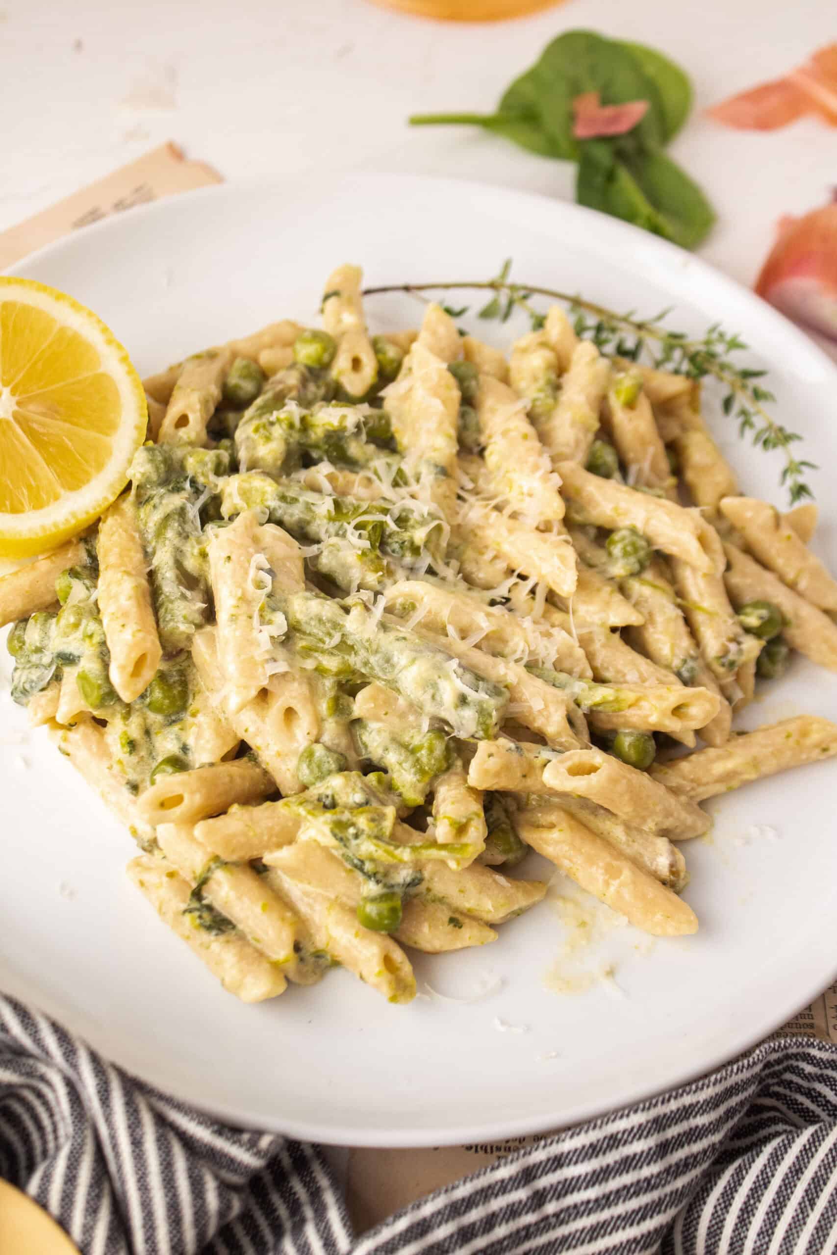 Penne pasta on a plate with asparagus, spinach, and peas with a slice of lemon.
