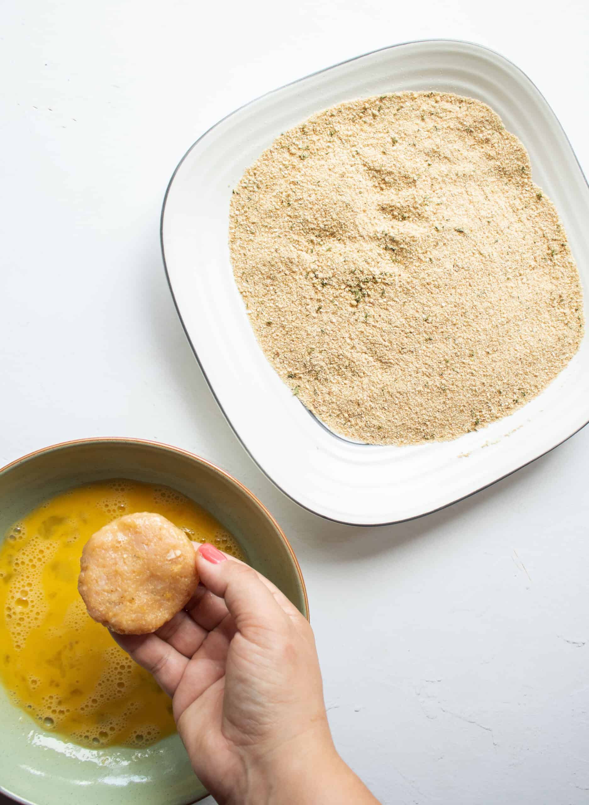 A hand dipping a chicken nugget patty into egg and then into breadcrumbs.