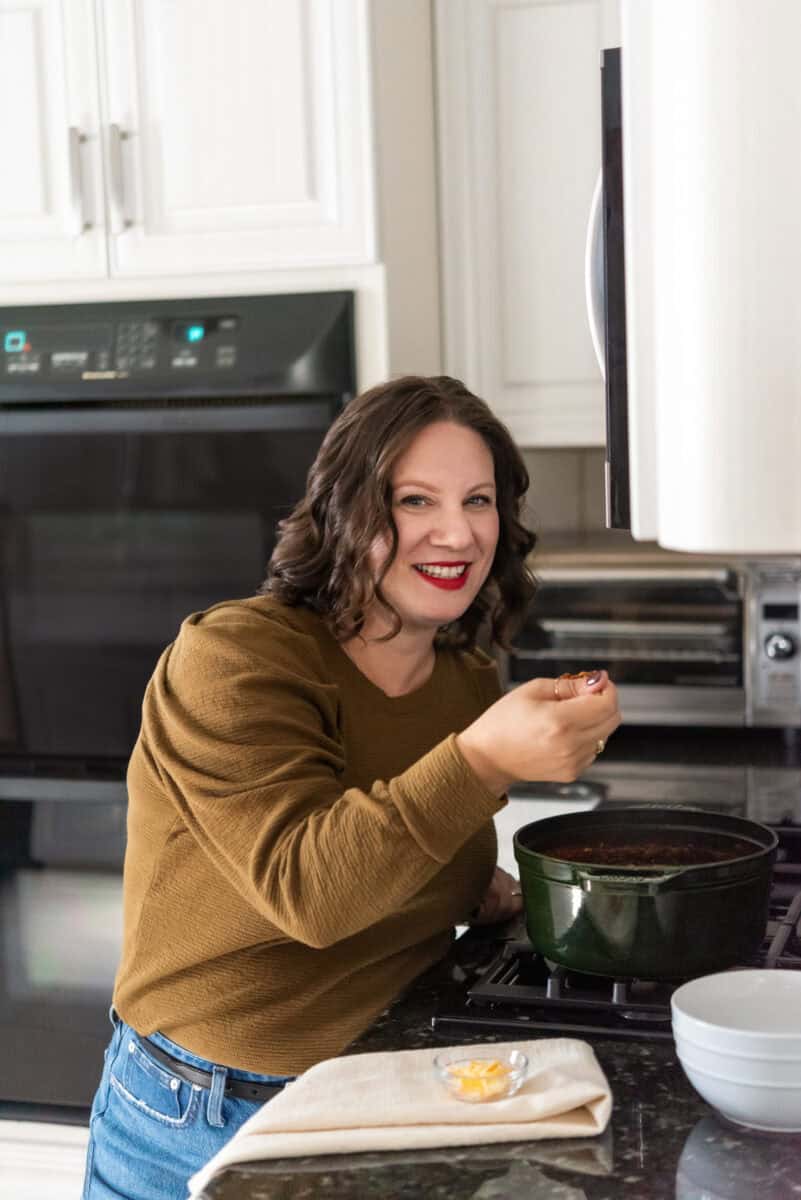 Morgan standing by her stove tasting chili from a green dutch oven on a wooden spoon.
