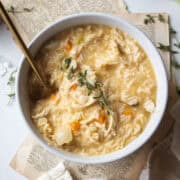 A large bowl of creamy chicken and rice soup garnished with fresh thyme surrounded by celery, garlic cloves, with a spoon in the soup.