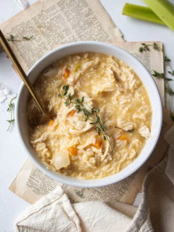 A large bowl of creamy chicken and rice soup garnished with fresh thyme surrounded by celery, garlic cloves, with a spoon in the soup.