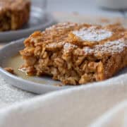 Close up of a plate on old paper with a slice of pumpkin baked oatmeal topped with maple syrup and powdered sugar.