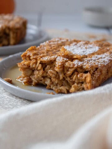 Close up of a plate on old paper with a slice of pumpkin baked oatmeal topped with maple syrup and powdered sugar.