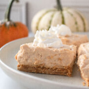 Close up of the profile of a pumpkin cheesecake bar on a plate with whipped cream and pumpkins in the background.