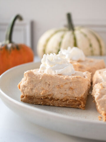 Close up of the profile of a pumpkin cheesecake bar on a plate with whipped cream and pumpkins in the background.