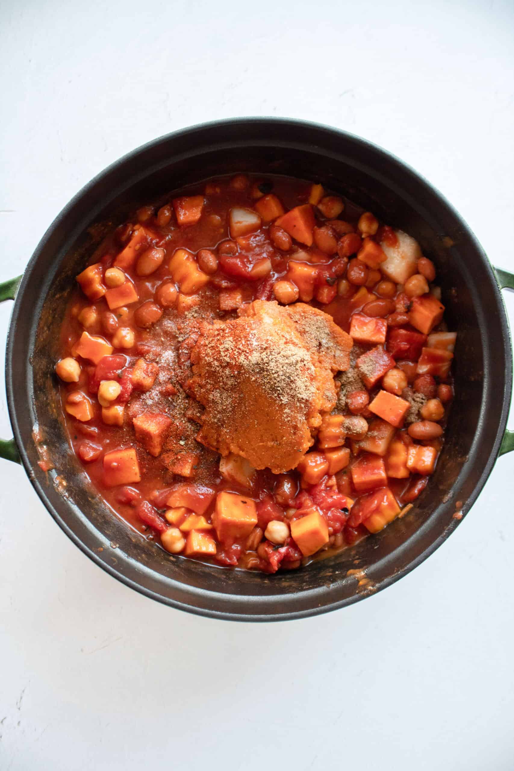 Pumpkin and seasoning on top of tomato sauce and diced tomatoes added to a dutch oven with butternut squash, sweet potato, onion, garlic, garbanzo beans.