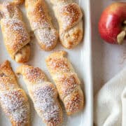 The corner of apple pie crescent rolls on a white platter with powdered sugar and an apple shown.