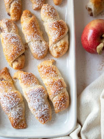 The corner of apple pie crescent rolls on a white platter with powdered sugar and an apple shown.