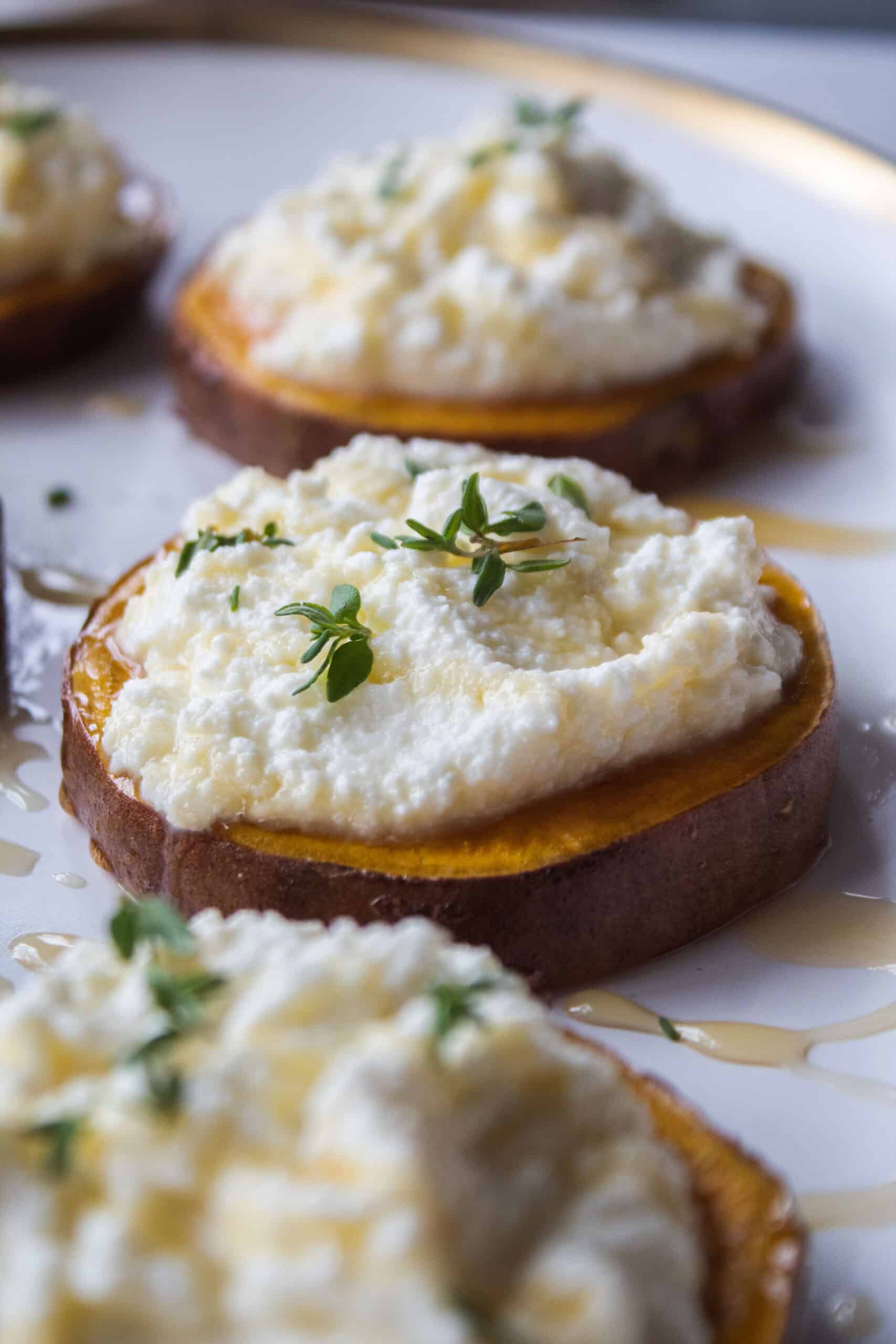 Close up of a sweet potato round topped with ricotta, thyme, and honey on a plate surrounded by other sweet potato rounds.