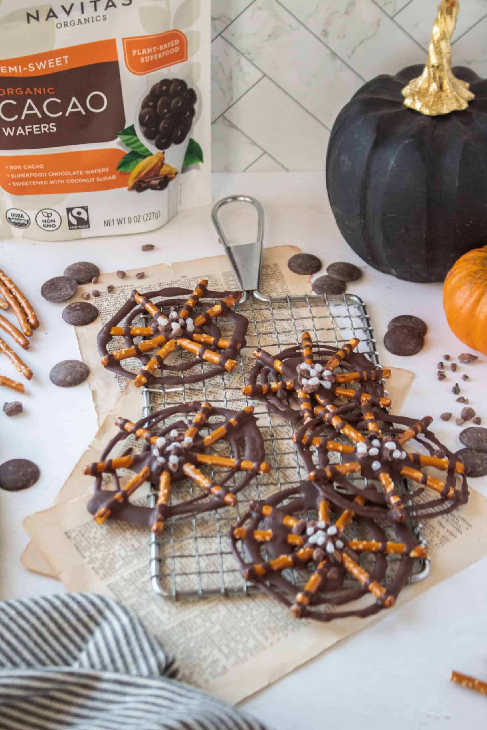 Five chocolate pretzel spider webs on a cooling rack surrounded by chocolate wafers, cacao nibs, pumpkins, and pretzels.