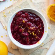 Homemade cranberry sauce in a white ramekin topped with fresh orange zest surrounded by oranges.