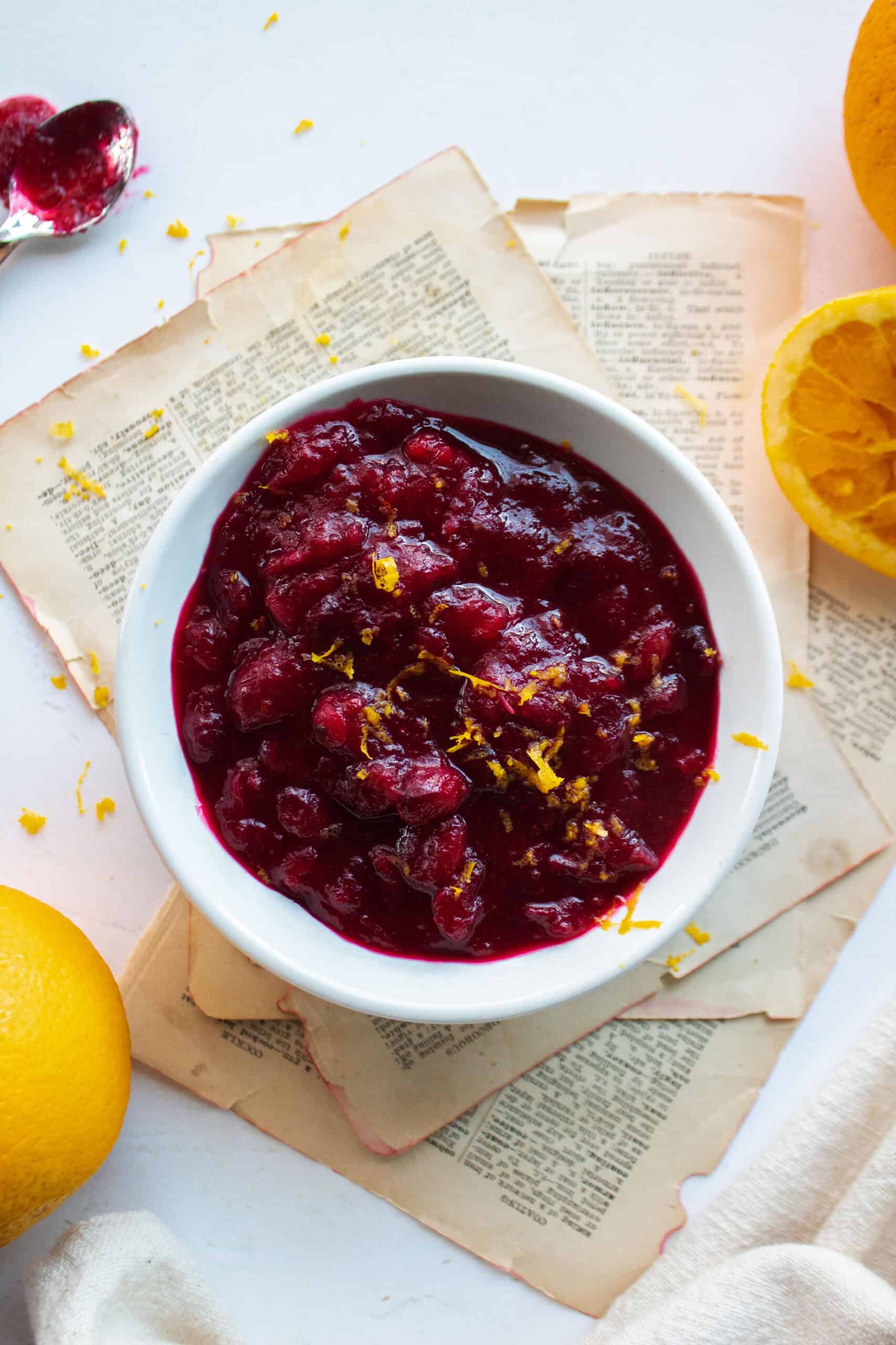 Homemade cranberry sauce in a white ramekin topped with fresh orange zest surrounded by oranges.