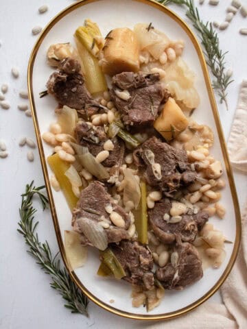 Braised lamb loin chops with white beans on a platter with braised vegetables and rosemary.