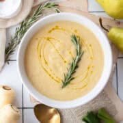 Pear and roasted parsnip soup in a white bowl garnished with rosemary and a drizzle of olive oil.