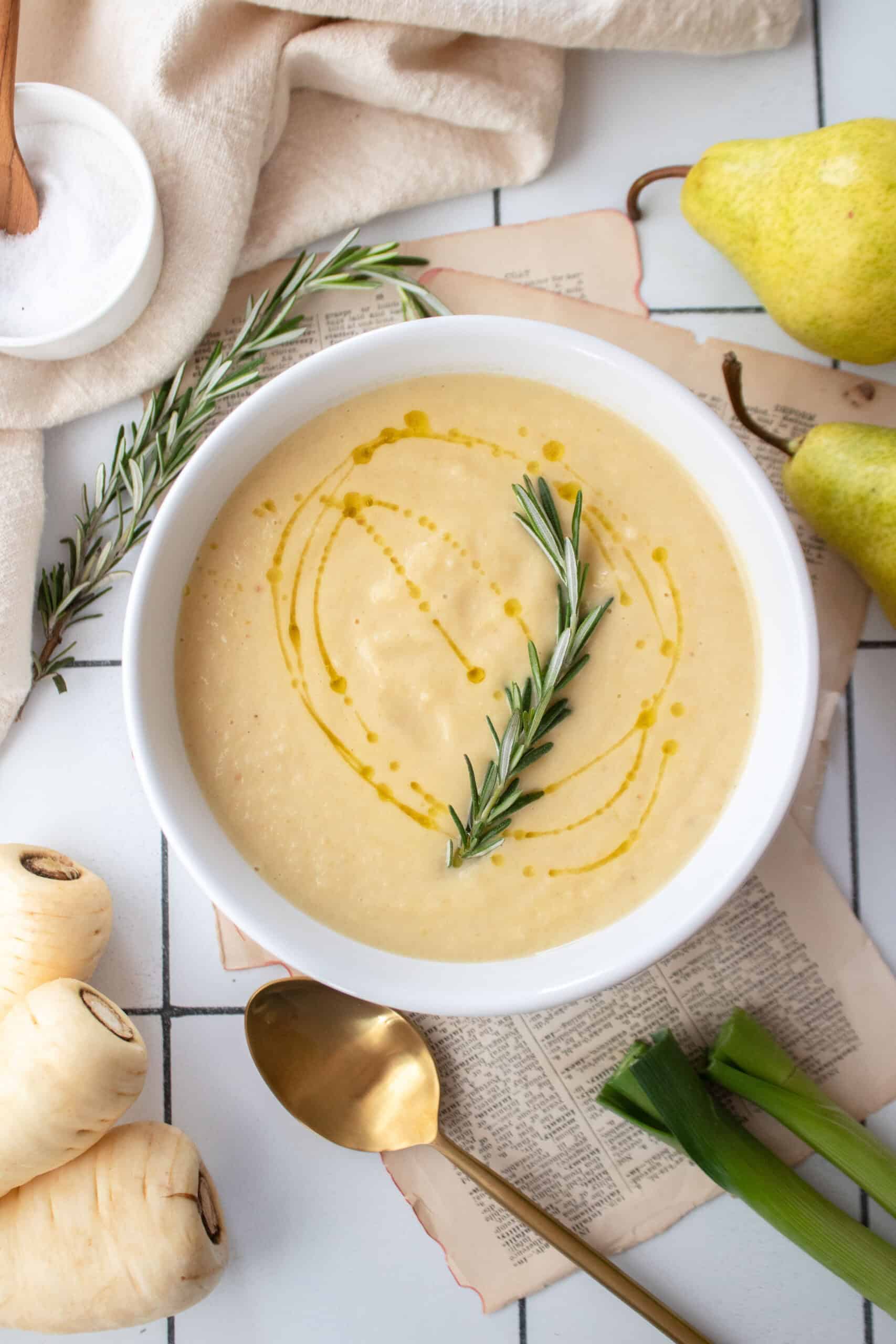 Pear and roasted parsnip soup in a white bowl garnished with rosemary and a drizzle of olive oil.
