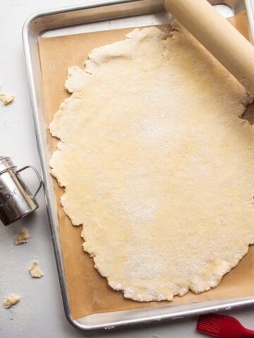 Galette pastry dough rolled out into an oval shape on a piece of parchment on a baking sheet with dusted flour and a rolling pin.