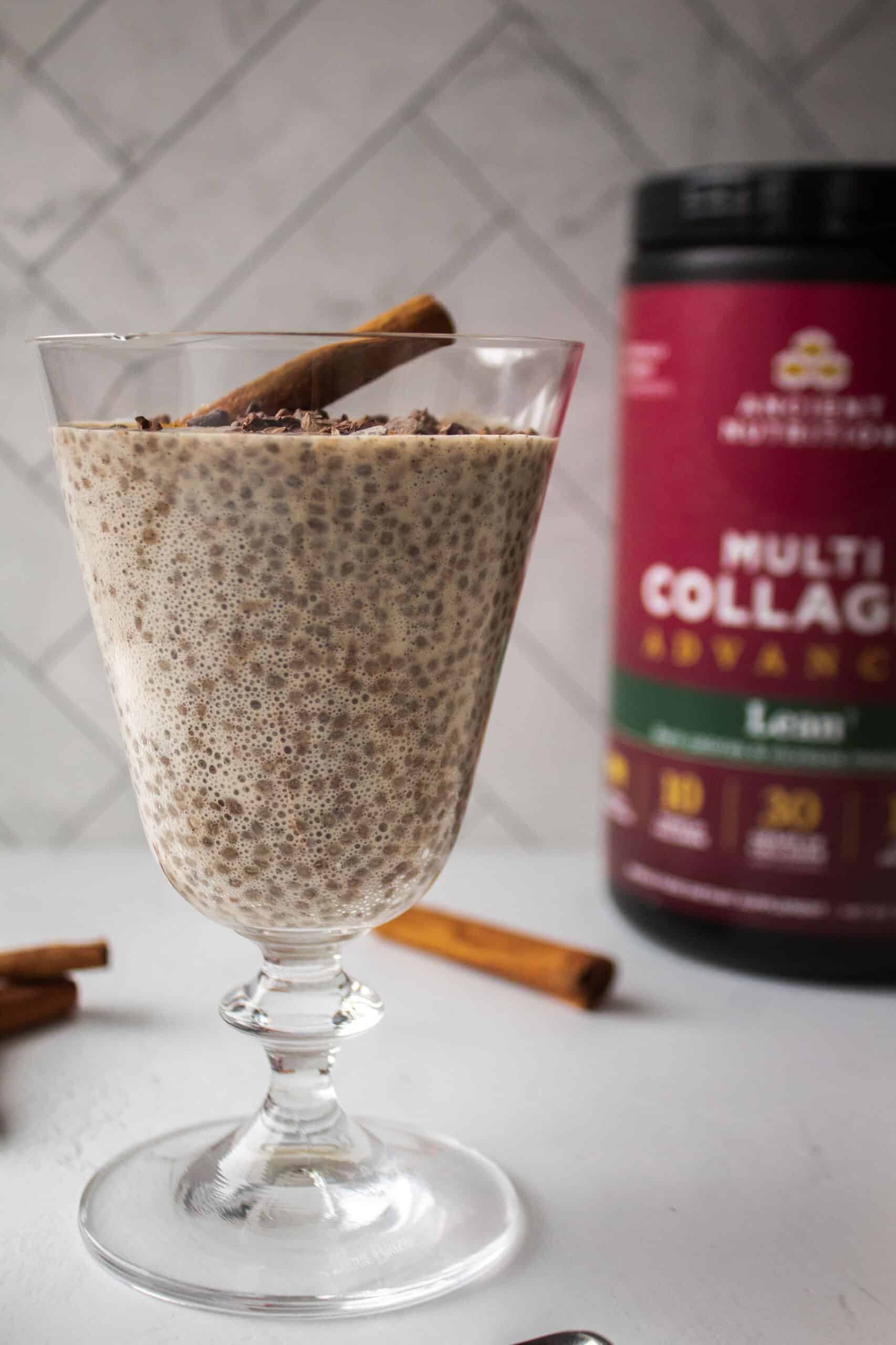 Cinnamon chia seed pudding with a scoop of Multi Collagen Advanced Lean in a glass topped with cacao nibs and a cinnamon stick on a white board.