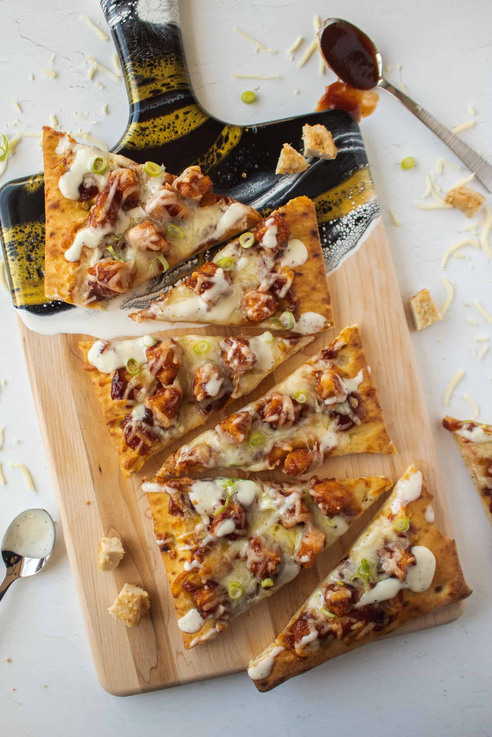 Pieces of a barbecue chicken flatbread pizza on a gold and wooden serving board topped with ranch dressing and scallions.