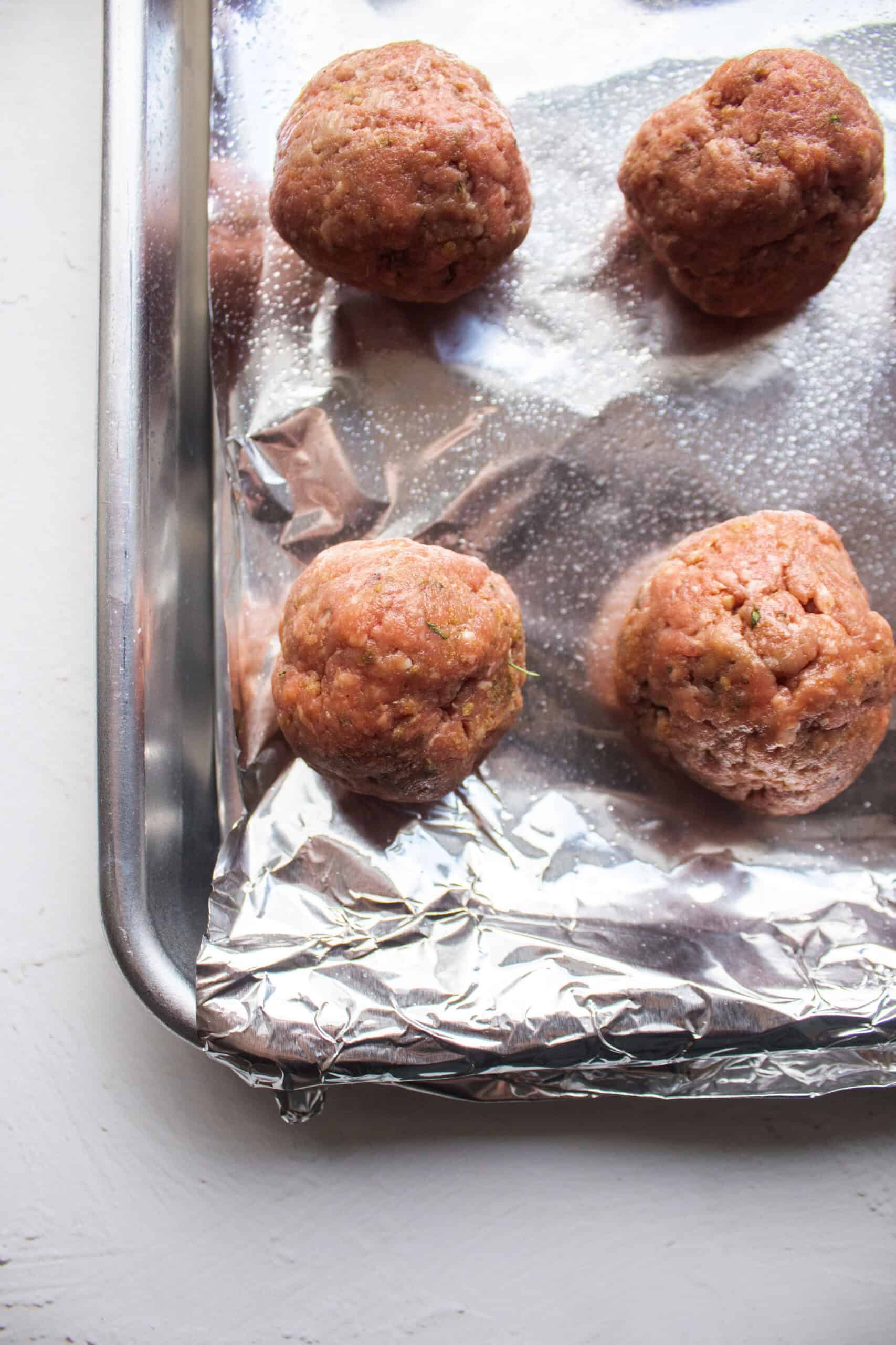Four mediterranean meatballs on a baking sheet lined with aluminum foil before baking.