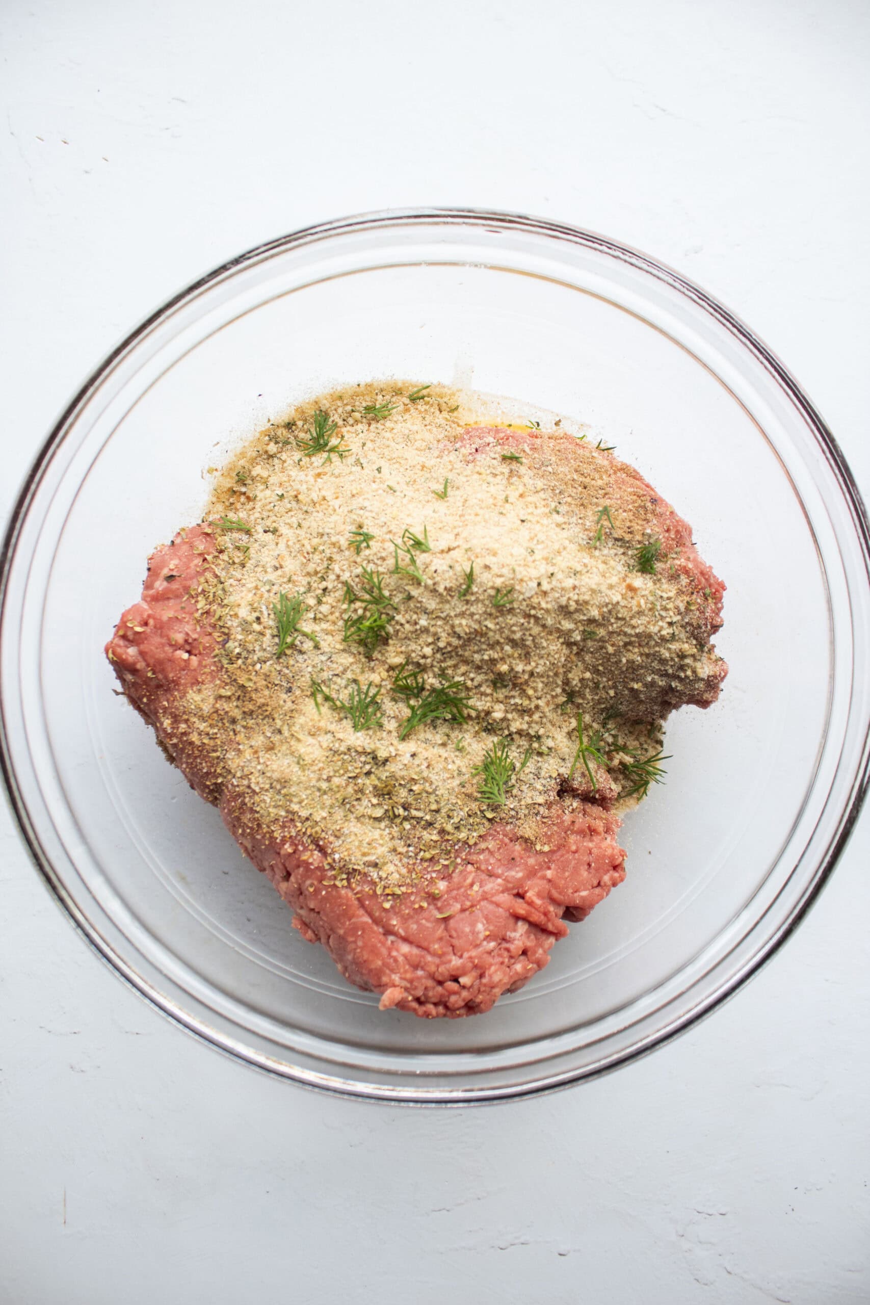 Ground beef, egg, worcestershire sauce, seasonings, breadcrumbs, and ill sprigs in a large glass mixing bowl.