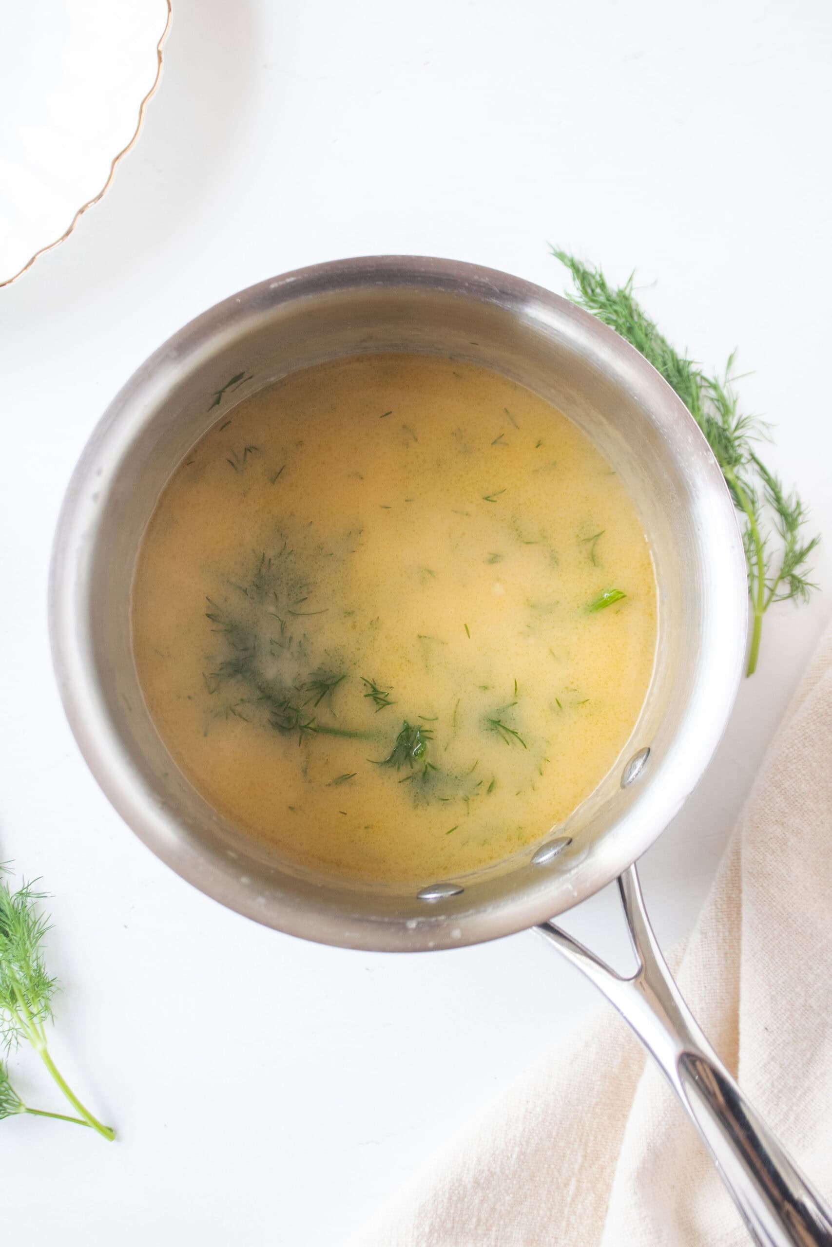 Melted butter mixed together with flour, beef broth, salt, dill sprigs to make a dill gravy simmering in a small saucepan.