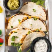 Six baked ground turkey tacos on a sheet pan topped with cilantro and surrounded by guacamole and sour cream.