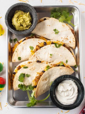 Six baked ground turkey tacos on a sheet pan topped with cilantro and surrounded by guacamole and sour cream.
