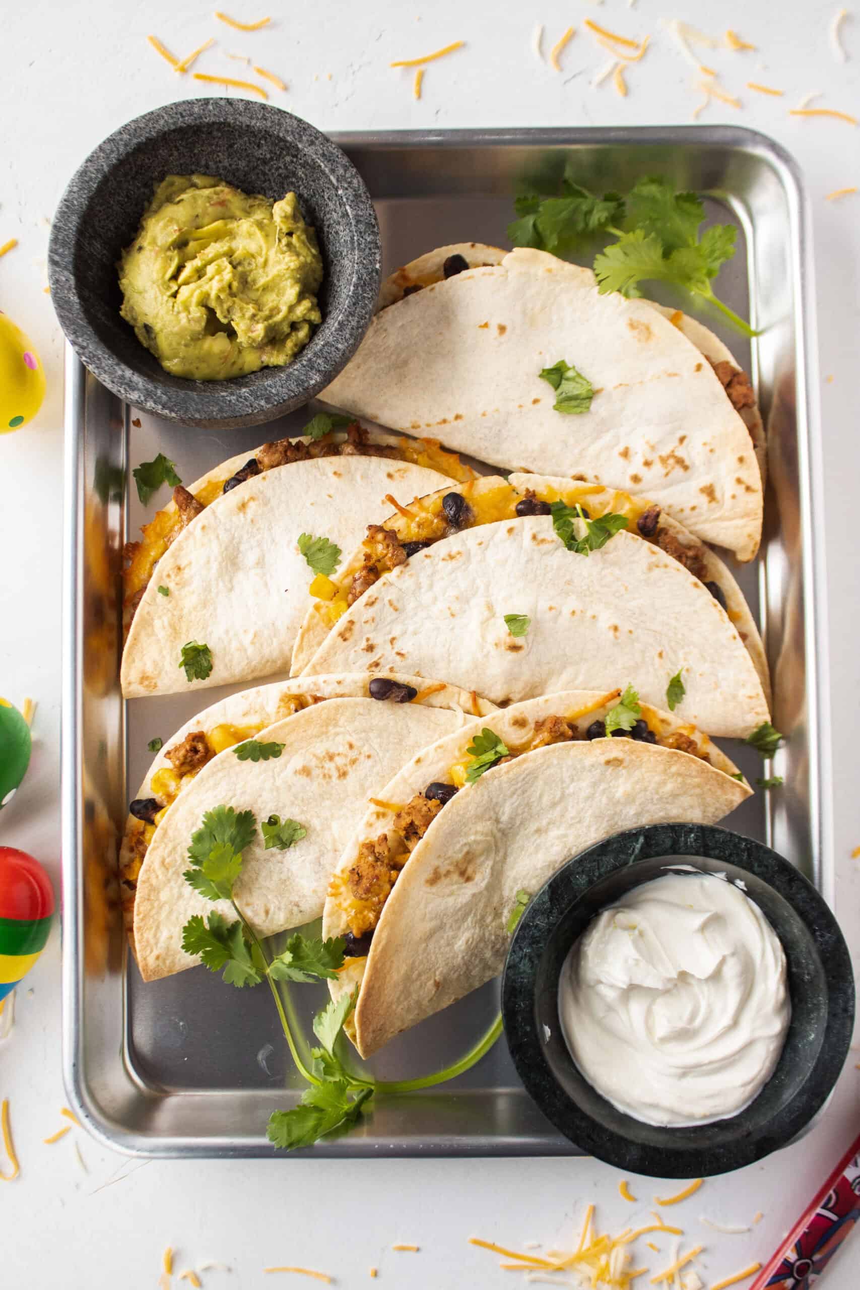 Six baked ground turkey tacos on a sheet pan topped with cilantro and surrounded by guacamole and sour cream.