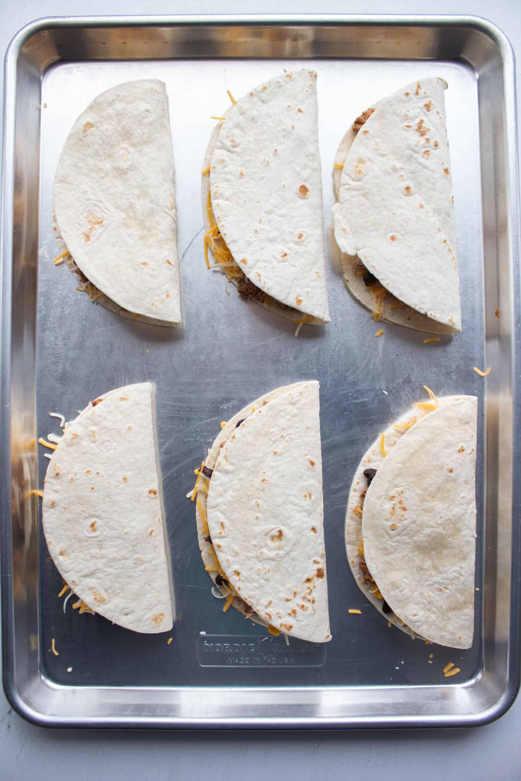 Six folded ground turkey tacos on a sheet pan before baking in the oven.