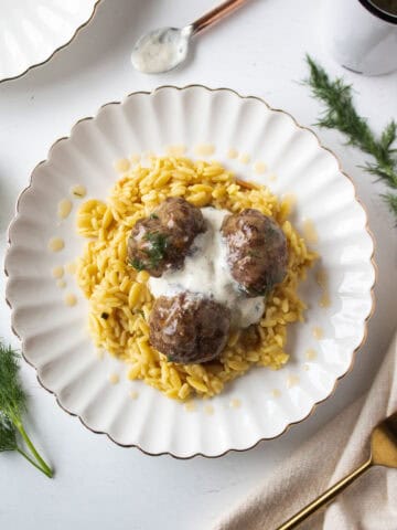 Three mediterranean meatballs on a plate with rice pilaf drizzled with dill gravy and a dollop of tzatziki in the middle.
