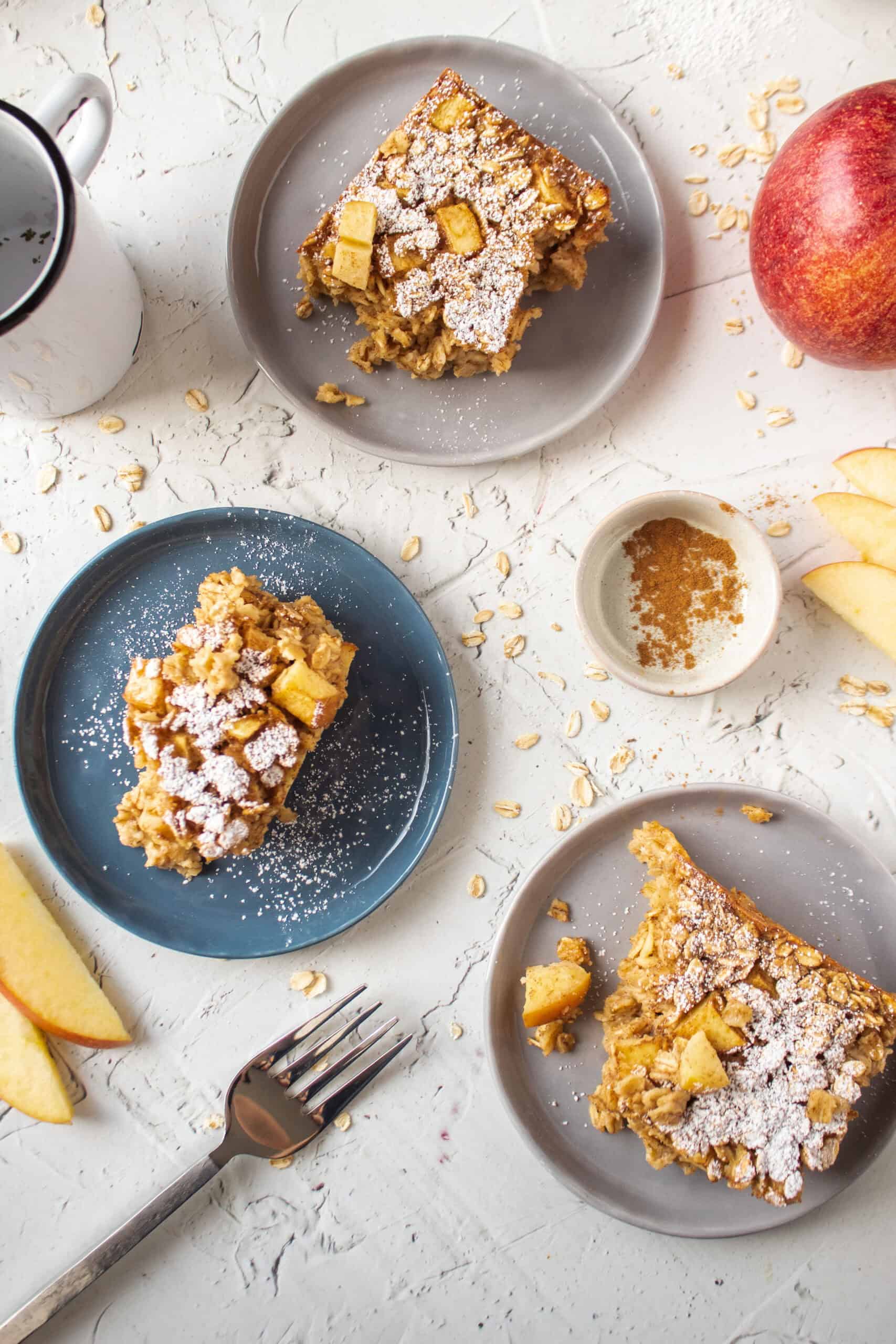 Three slices of apple cinnamon baked oatmeal on three small appetizer plates topped with powdered sugar surrounded by sliced apple and oats.