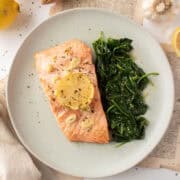 Two filets of salmon topped with a lemon, butter, garlic sauce on a plate with sauteed spinach surrounded by lemons and a napkin.